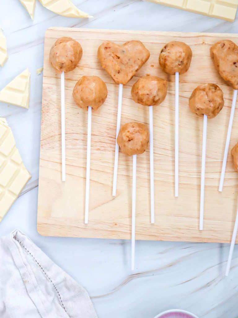Cake pops formed and on sticks, ready to be coated in chocolate. They are all lined up and lying on a wooden cutting tray.