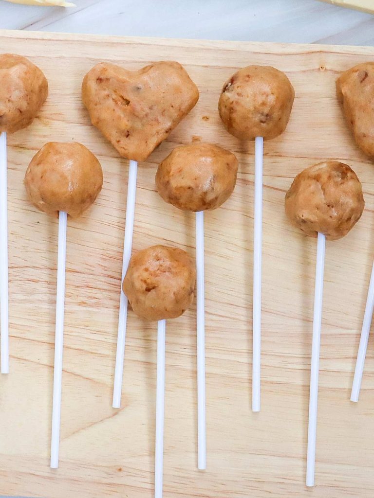 A close up shot of formed cake pops that are on sticks lying on a wooden cutting board, ready to be dipped into chocolate