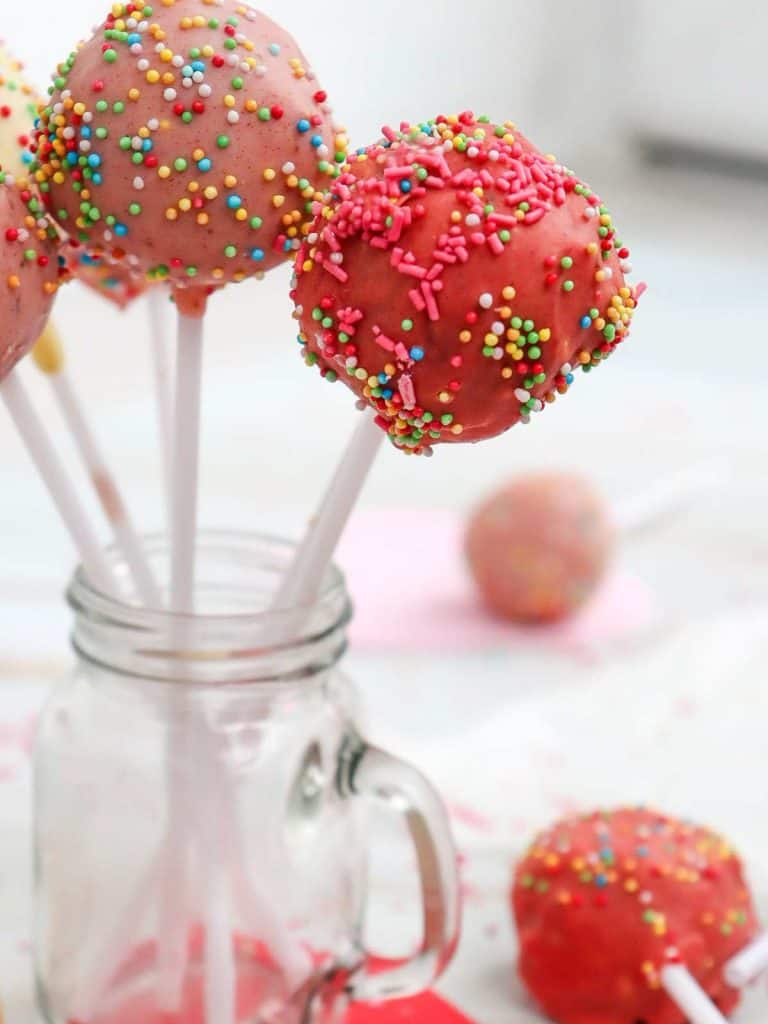 A close up of the finished cake pops in multiple shades of pink with different colored sprinkles decorating the outside of the cake pops and they are sitting upright in a jar