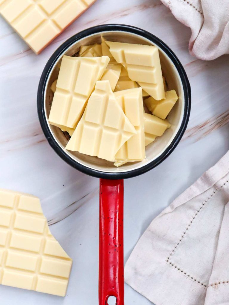 A white chocolate bar broken into pieces and put in a small red sauce pan to melt on the stove