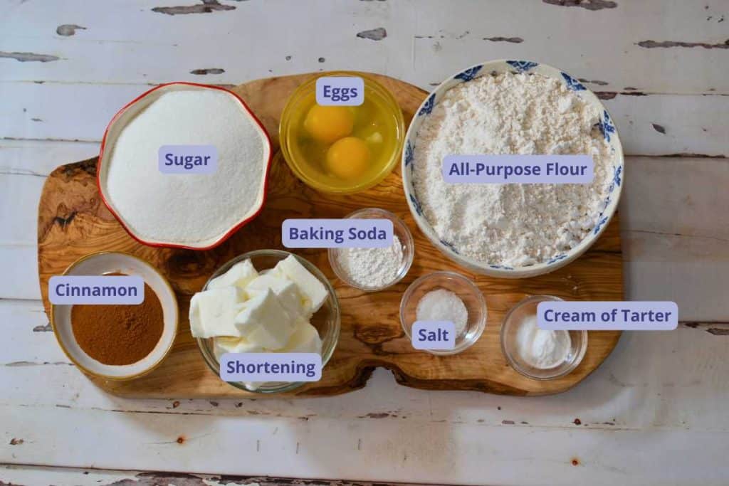 Ingredients for Snickerdoodle cookies in bowls on a wooden board, including flour, cream of tarter, salt, baking soda, eggs, sugar, shortening and cinnamon.