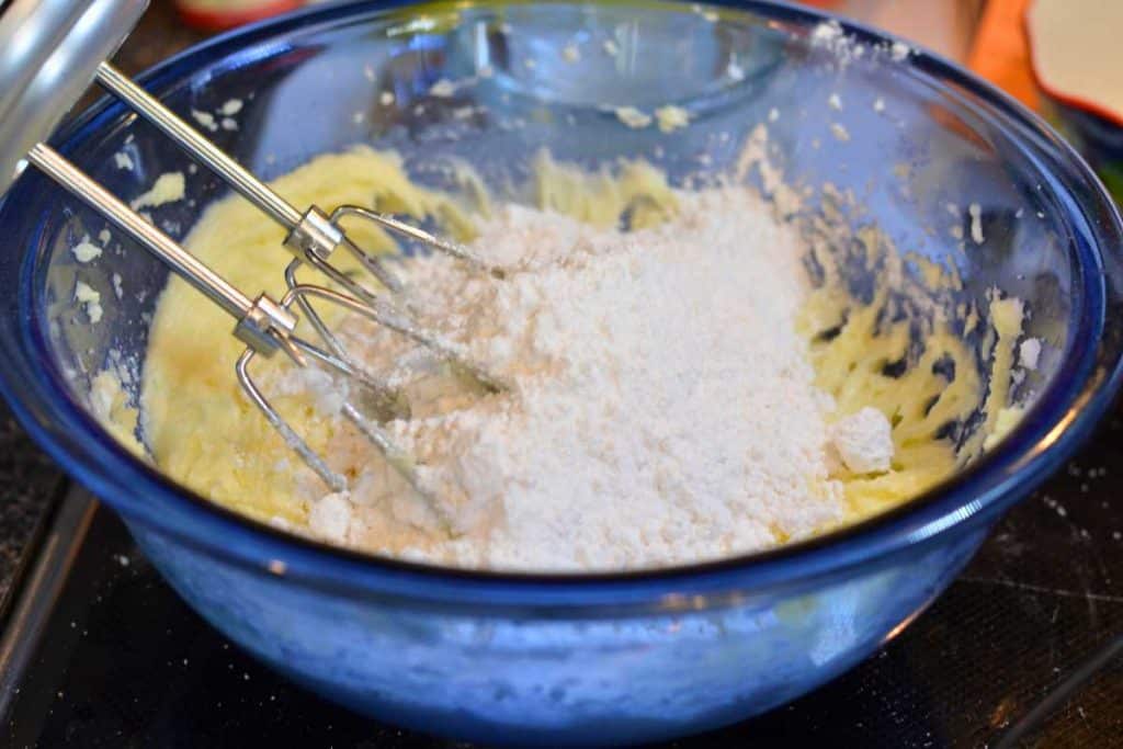 A blue glass mixing bowl with Snickerdoodle cookie dough with flour added to the bowl with an electric mixer.