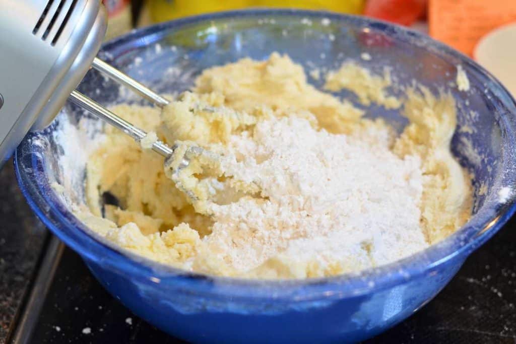 Second addition of flour to snickerdoodle cookie dough in a blue glass bowl with an electric hand mixer.