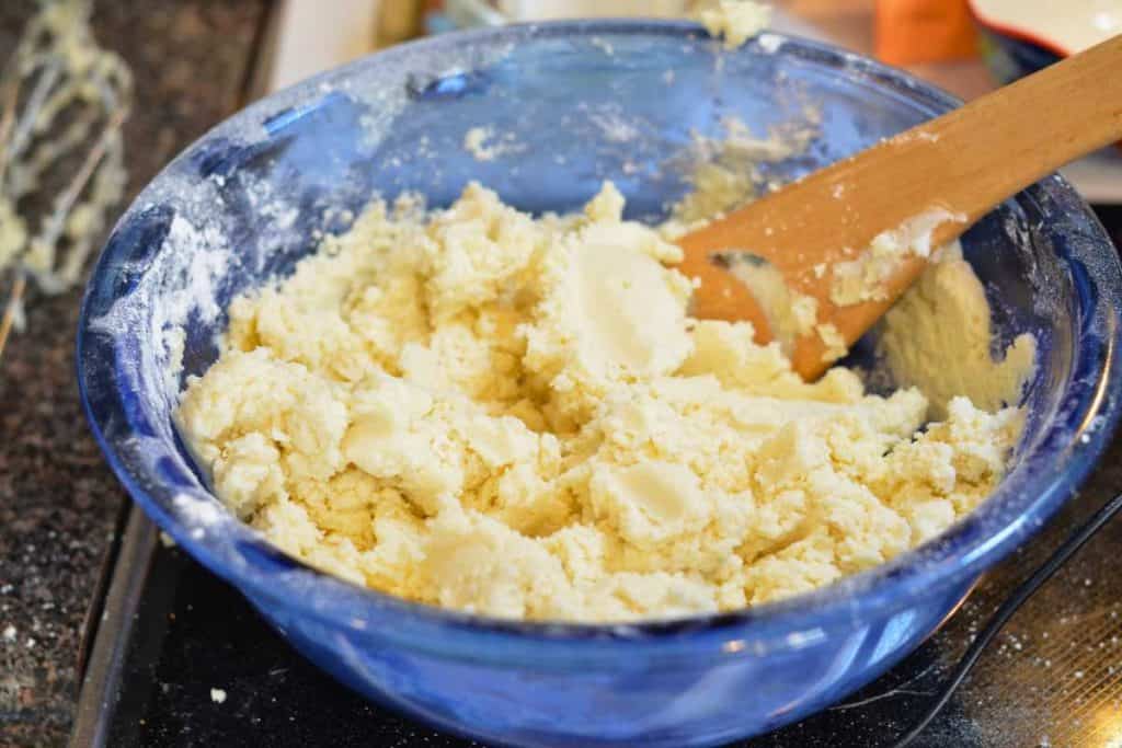A wooden spoon in the snickerdoodle cookie dough finishing up the mixing of the dough. The dough is in a blue glass bowl.