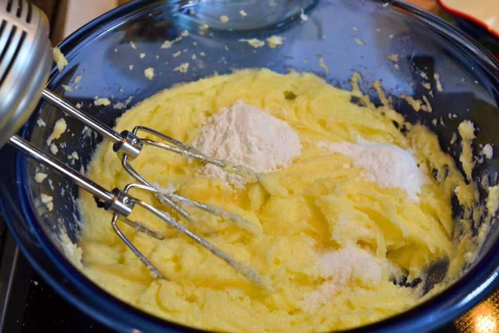 Salt, baking soda and cream of tarter added to the Snickerdoodle cookie dough in a blue glass bowl with an electric mixer in the corner of the bowl.
