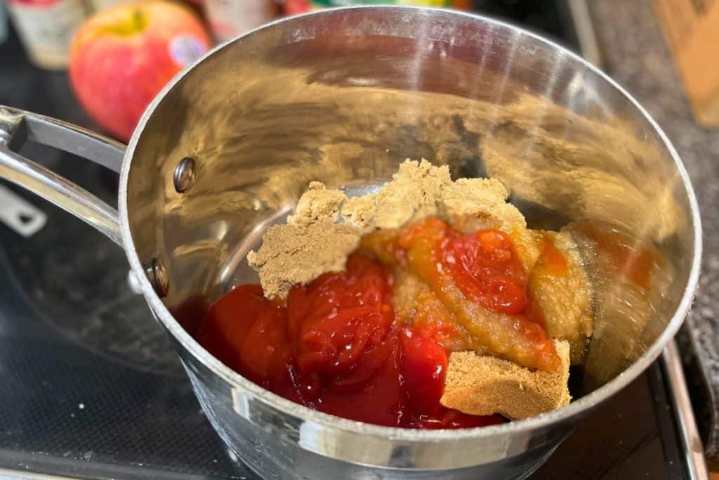 A saucepan with the beginning ingredients of apple bbq sauce, including brown sugar, ketchup and applesauce.