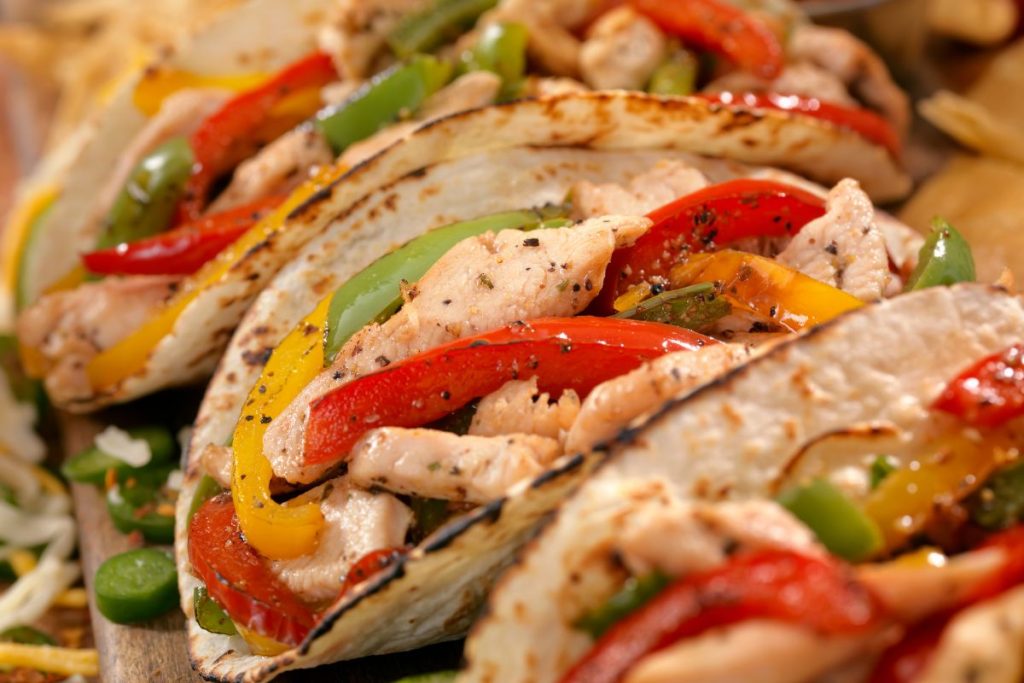 A close up of chicken fajitas all assembled and ready to eat in flour toritillas lined up on a wooden cutting board.