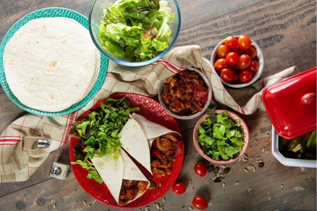 A fajita dinner spread that includes a plate of tortillas, a bowl of salad, the fajita mixture in a bowl, tomatoes ina bowl, and a plate of fajitas all made up and ready to eat wrapped in a flour tortilla.