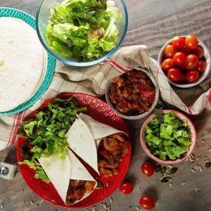 A chicken fajita dinner spread from above with a plate of tortillas, a bowl of salad, several bowls of the fajita fixings and a plate with 2 flour tortillas stuffed with fajita mixture.