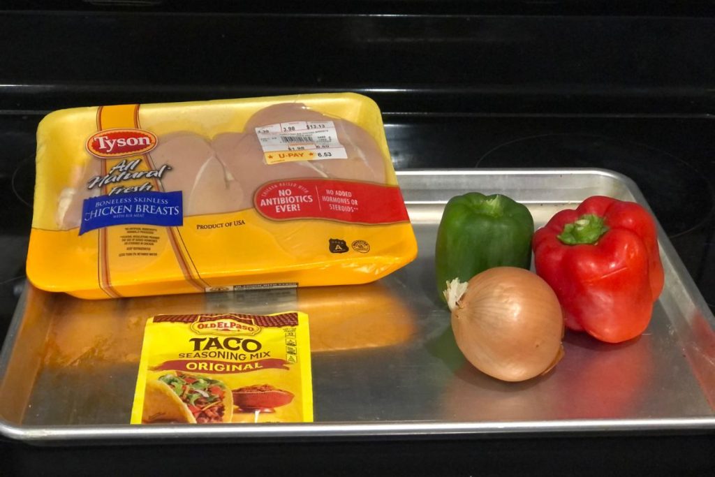 The ingredients for sheet pan chicken fajitas which include a package of boneless chicken breasts, a green bell pepper, a red bell pepper, a yellow onion and a taco seasoning pack. All of this is on a sheet pan.