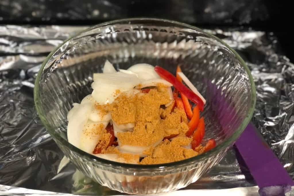 The fajita vegetables in a glass bowl with the seasoning sprinkled on top of them ready to mix. Next to the bowl is a purple knife.