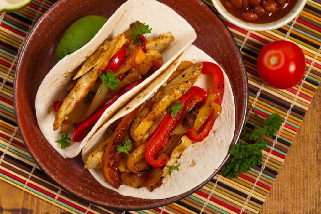 Finished chicken fajitas on a plate with flour tortiallsa dn beans and a slice of lime.