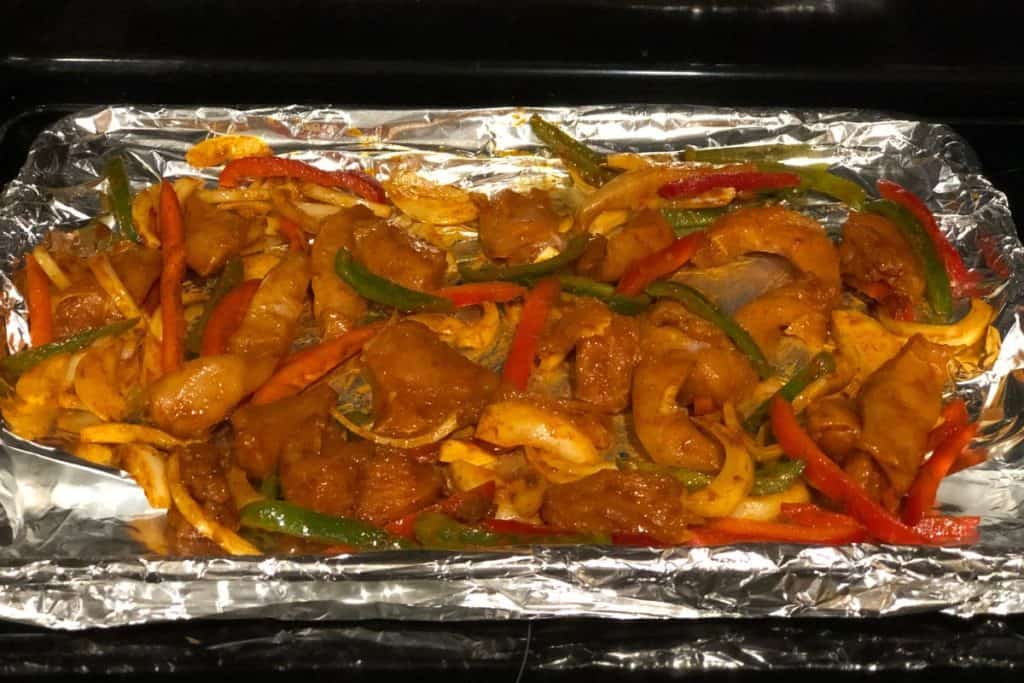 A sheet pan covered in aluminum foil with raw fajita ingredients on it including chicken, onions and red and green bell peppers. All are covered in a marinade seasoning and are ready to roast in the oven.