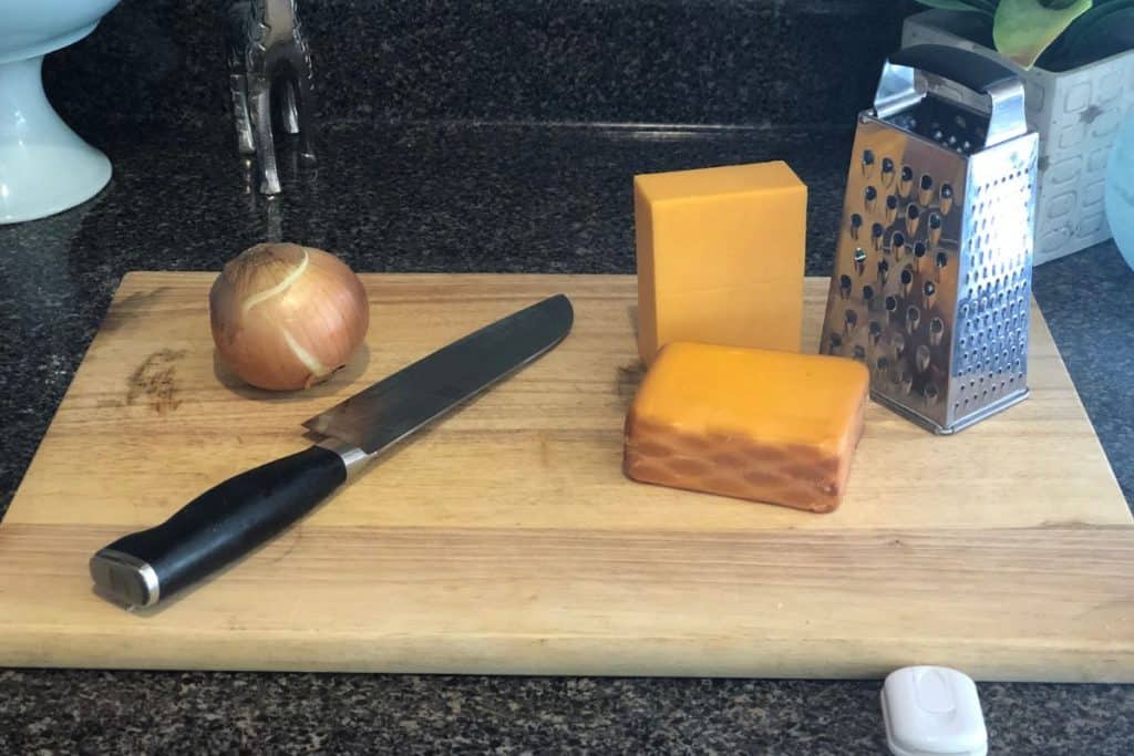 Two blocks of cheese and a yellow onion on a cutting board with a box grater and a santoku knife.