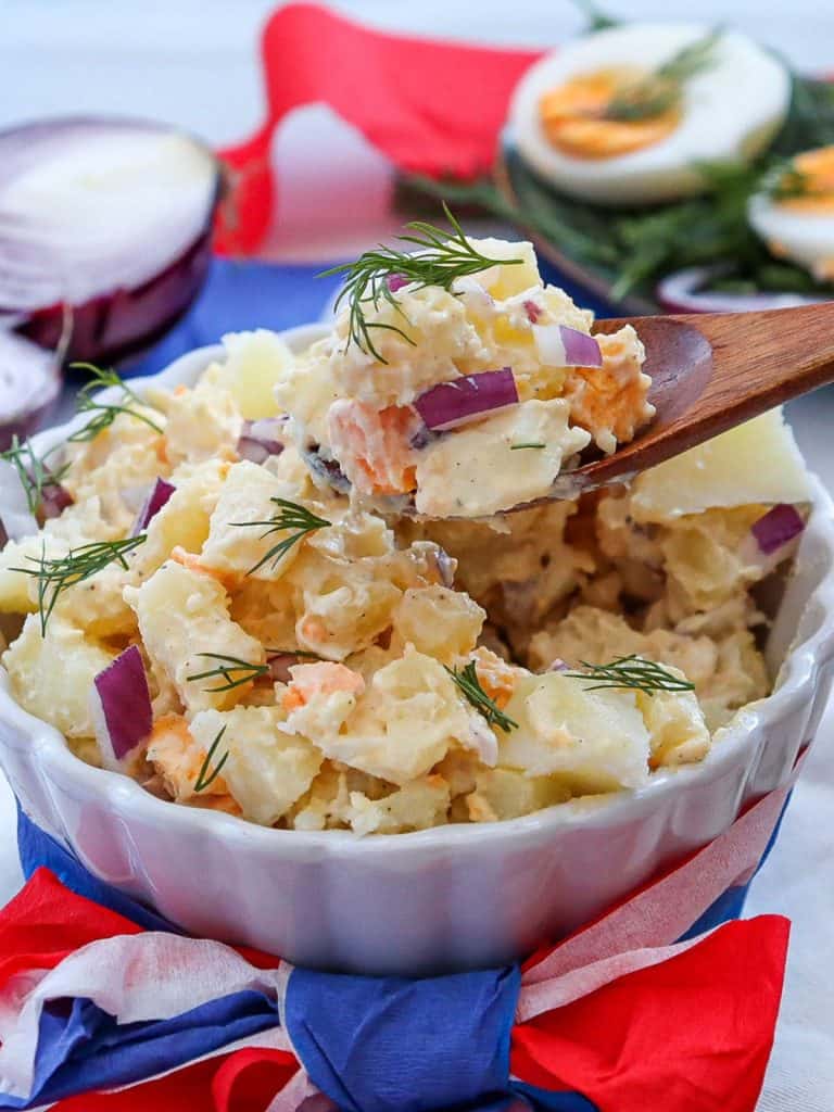 white ceramic bowl of potato salad with red, white and blue ribbon wrapped around the bowl with a wooden spoon taking a spoonful of potato salad