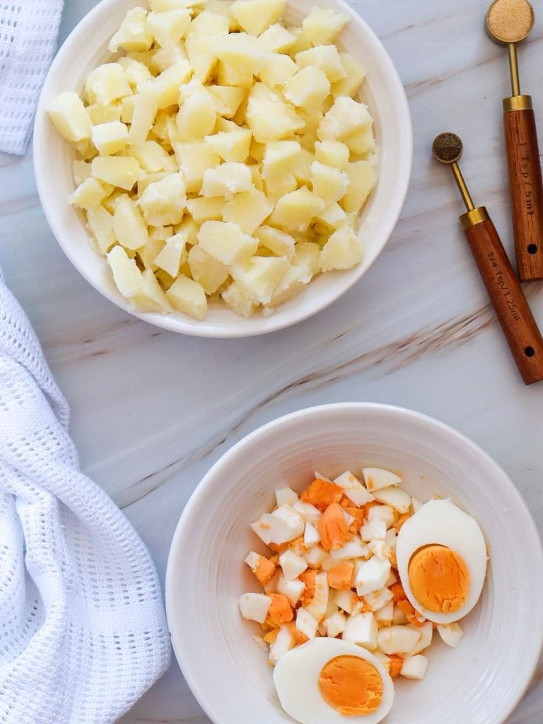 a white bowl of cubes of cooked potato and a white bowl of chopped hard boiled eggs