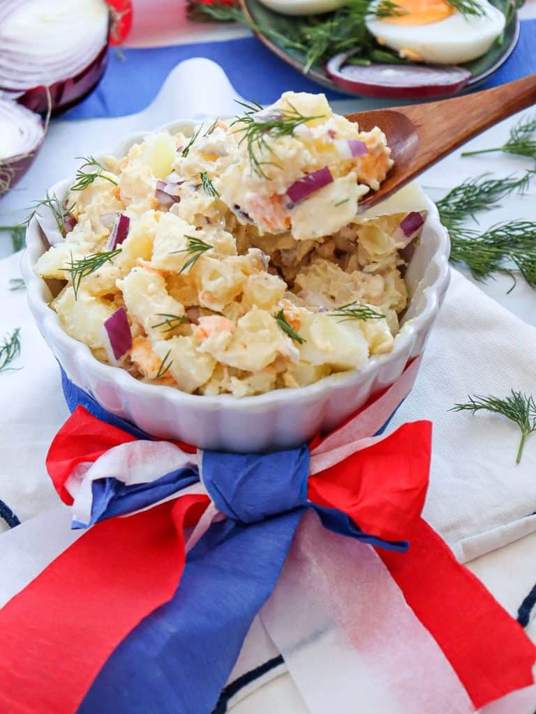 white ceramic bowl of potato salad with red, white and blue ribbon wrapped around the bowl with a wooden spoon taking a spoonful of potato salad