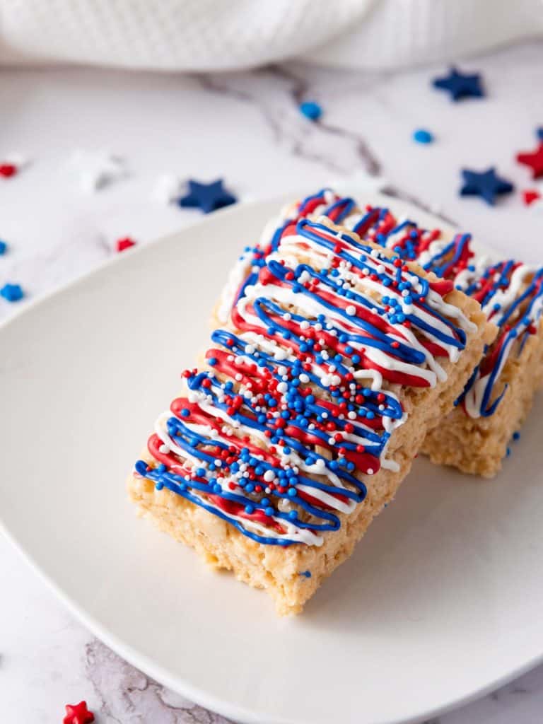 A plate with two rice krispy treats on it and they are glazed with red, white and blue icing and sprinkles