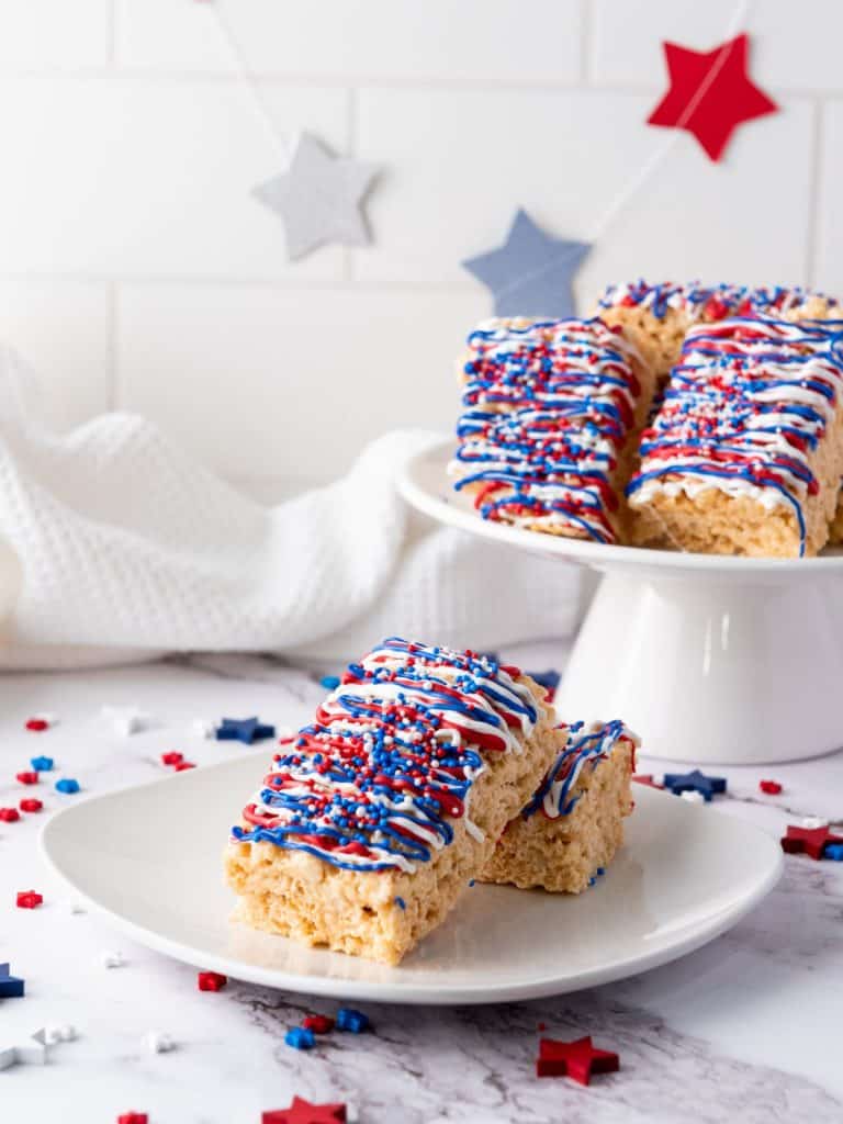 A white cake pedestal with patriotic rice krispie treats and a white plate with two rice krispie treats on it