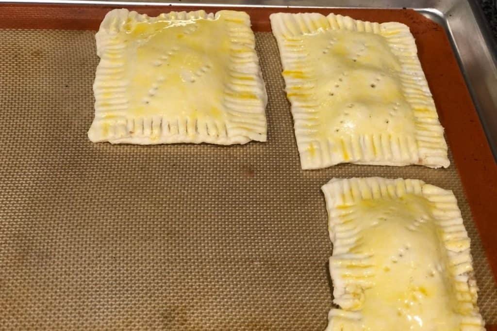 Ready to go into the oven pop tarts sitting on a baking sheet with egg wash and holes poked in the top of the pastry crust for ventilation.