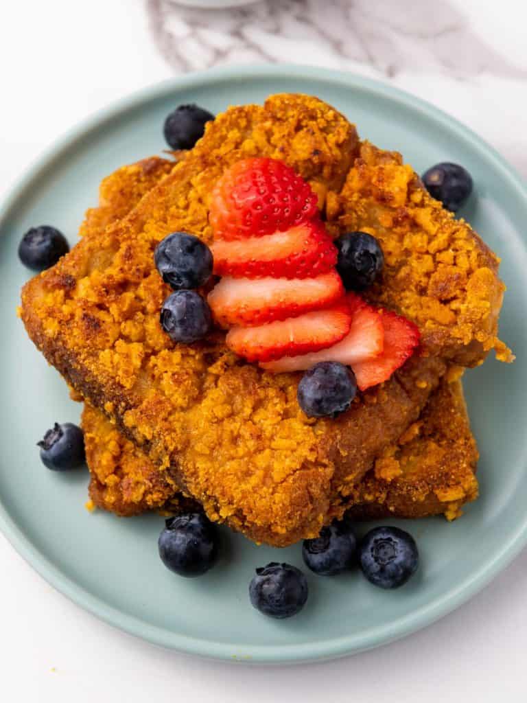 A close up of finished Captain Crunch French Toast served on a blue plate with blueberries and strawberries.