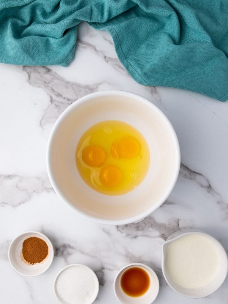 3 cracked eggs in a white bowl with other wet ingredients surrounding the bowl that are heavy cream, vanilla extract, sugar and cinnamon.