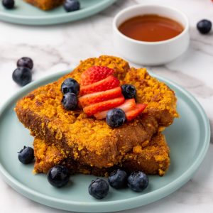 A close up of cooked Captain Crunch French Toast with a blue plate and served with syrup, blueberries and strawberries.