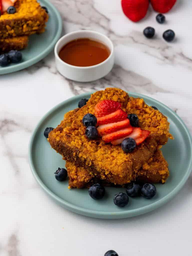 An side view of finished Captain Crunch French Toast served with syrup, blueberries and sliced strawberries.