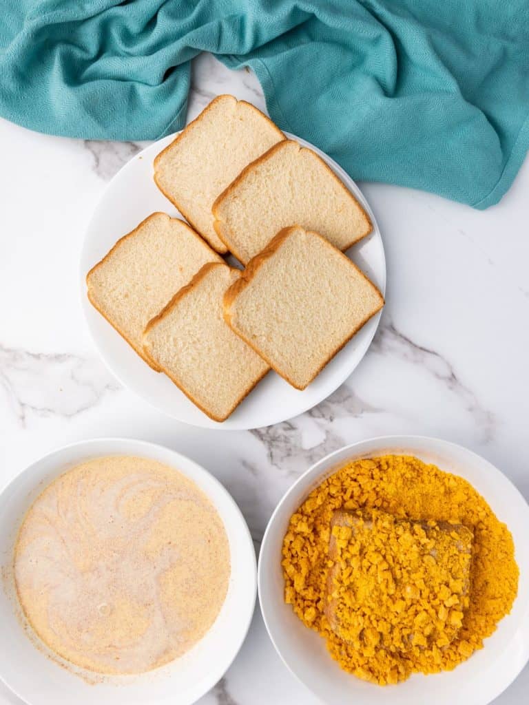 Breading station for Captain Crunch French Toast that includes a platter of white bread, the egg mixture in a bowl, and a bowl of crushed Captain Crunch cereal.