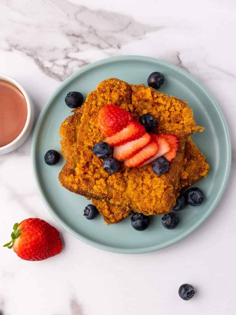 Above view of Captain Crunch French Toast on a blue plate topped with blueberries and a sliced strawberries, with syrup on the side in a white ramikin.