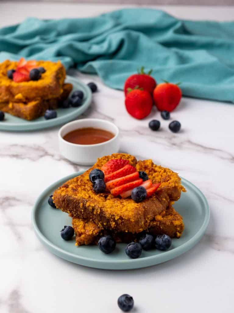 Captain Crunch French Toast on a blue plate topped with blueberries and a sliced strawberries, with syrup on the side in a white ramikin.