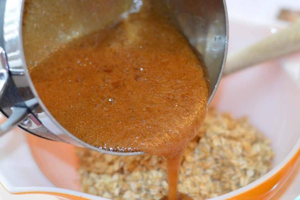 Pouring the boiling brown sugar mixture over the toasted oats in a large bowl with a wooden spatula