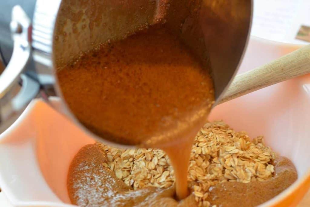 Pouring the boiling brown sugar mixture over the toasted oats in a large bowl with a wooden spatula