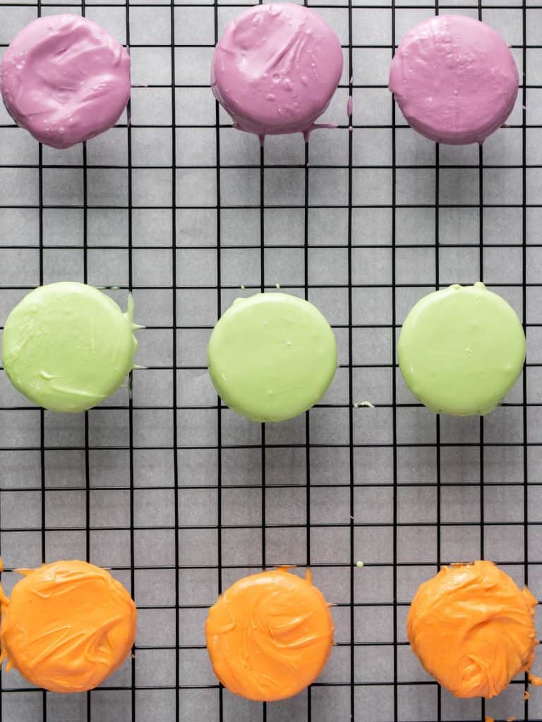 Oreos dipped in purple, green, and orange candy coating set on a wire rack to dry.