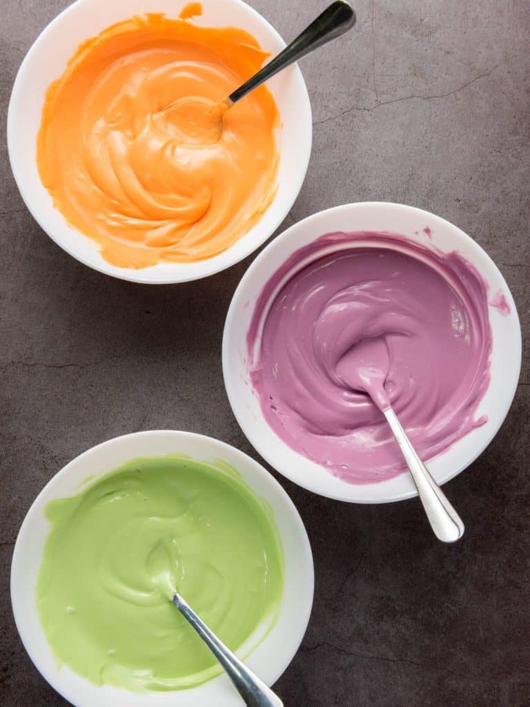 Three bowls of melted purple, green, and orange candy melts with spoons, ready for dipping Oreos.